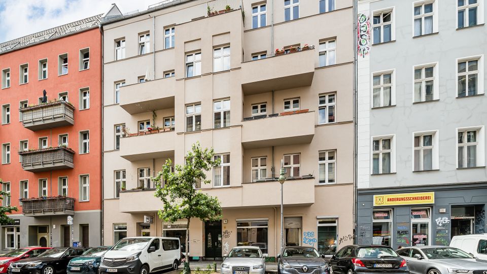 Straßenansicht mehrerer mehrstöckiger Wohnhäuser mit hellen Fassaden und gestaffelten Balkonen, teils mit Blumenkästen. Im Vordergrund stehen geparkte Autos, ein kleiner Straßenbaum und eine Laterne; im Erdgeschoss sind Ladenfronten mit Schaufenstern zu sehen.