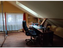 Dachzimmer mit einem Schreibtisch und Bürostuhl in einer Ecke unter einer schrägen Decke. Der Raum hat Holzfußboden, ein Fenster mit weißen Gardinen und orangenen Vorhängen sowie sichtbare Holzträger.