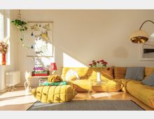 Wohnzimmer mit einer großen, gelben Ecksofa-Landschaft, einem passenden Polsterhocker und einem transparenten Couchtisch. Der Raum ist hell, mit Sonneneinstrahlung, dekoriert mit Pflanzen, farbigen Kissen, Büchern und einer Stehlampe mit goldenem Lampenschirm.