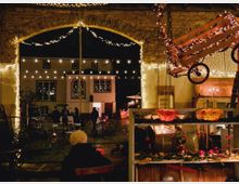 Ein weihnachtlich geschmückter Innenhof mit stimmungsvoller Beleuchtung, einer Backsteinbogenöffnung und Lichterketten, die über den Platz gespannt sind. Im Vordergrund stehen ein Verkaufstresen mit Dekorationen und ein rustikaler hölzerner Wagen, der von Lichtern umwickelt ist, während Menschen sich an Tischen und Ständen versammeln.