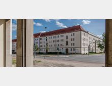 Straßenszene mit einem großen Wohngebäude im Stil der Nachkriegsarchitektur, hell verputzt mit roten Satteldächern und zahlreichen Fenstern. Im Vordergrund ist eine Verkehrsinsel mit Kreuzung, Fußgängerampeln und Straßenlaternen zu sehen.