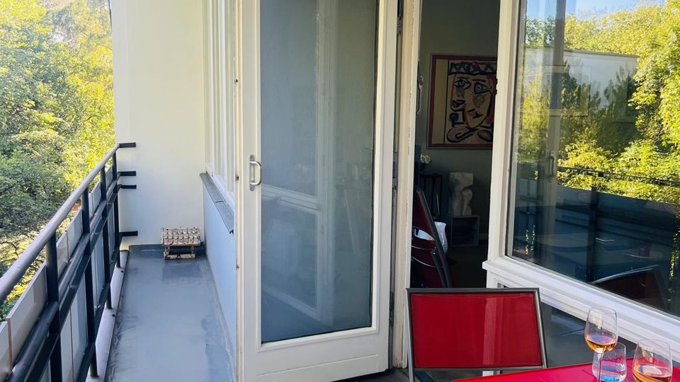 Schmaler Balkon mit grauem Boden und schwarzem Metallgeländer, angrenzend an eine Wohnung mit großen Fenstern und einer geöffneten Glastür. Rechts steht ein roter Tisch mit Stühlen und Sitzkissen; im Hintergrund ist eine grüne Baumlandschaft zu sehen.