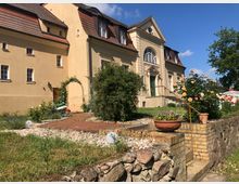 Gelbes, historisches Gebäude mit einer symmetrischen Fassade und einem großen, verzierten Eingang mit grüner Tür und rundem Fenster darüber. Im Vordergrund befindet sich ein gepflegter Garten mit Gehweg, Blumenbeeten und Rosenbüschen, umgeben von einer Natursteinmauer.