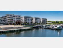 Moderne Wohnanlage mit mehreren vierstöckigen weißen Gebäuden und großen Balkonen, gelegen an einem Hafenbecken mit mehreren festgemachten Motorbooten. Im Vordergrund sind eine Promenade mit Treppenstufen und Bäumen sowie Wasserflächen zu sehen.