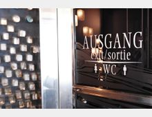 Schwarze Glastür mit der Aufschrift "AUSGANG exit/sortie WC" und Symbolen für Damen und Herren; rechts sind reflektierende Lichter erkennbar, links eine dekorative Wand mit quadratischen Kristallelementen.