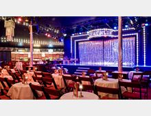 Innenraum eines Theaters oder Varietés mit Tischen und Stühlen, die auf eine Bühne mit glitzerndem Vorhang ausgerichtet sind. Die Bühne ist von Lichtern umrahmt, und im Hintergrund befindet sich eine beleuchtete Bar mit Regalen voller Flaschen.