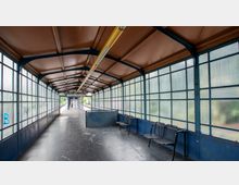 Überdachter Wartebereich einer Bahnstation mit einer schmalen Passage. Die Wände bestehen aus Glasfenstern mit Metallrahmen, der Boden hat eine dunkle Gummibeschichtung, und es gibt Metallbänke auf der rechten Seite.