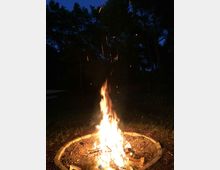 Ein Lagerfeuer in einer kreisförmigen, mit Steinen eingefassten Feuerstelle, umgeben von einer dunklen Waldlandschaft bei Nacht. Funken steigen vom Feuer in den dunklen Himmel auf.