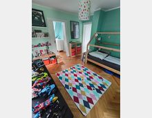 Kinderzimmer mit mintgrünen Wänden, einem Hochbett aus Holz auf der rechten Seite und einem bunten Teppich mit Rautenmuster in der Mitte. Links stehen Regale und ein Tisch mit Spielzeug, im Hintergrund führen zwei Türen zu anderen Räumen.