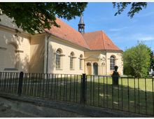 Eine beige Kirche im barocken Stil mit roten Ziegeldächern, halbkreisförmigen Fenstern und einem kleinen Dachreiter. Im Vordergrund befindet sich ein Metallzaun, dahinter eine Grünfläche mit Bäumen und einer Büste.