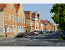 Straßenszene in einer Altstadt mit historischen Backsteinhäusern, die geschmückte Giebel und weiße Fensterläden haben. Auf der Straße stehen geparkte Autos, und im Vordergrund ist ein Fußgänger zu sehen; rechts säumen Bäume den Straßenrand.