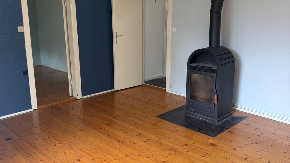 Leerer Wohnraum mit breiten Holzdielen und blau-weiß gestrichenen Wänden, zwei Türöffnungen führen in angrenzende Räume. Rechts steht ein schwarzer Kaminofen mit Ofenrohr unter der Decke und einer Metallbodenplatte.