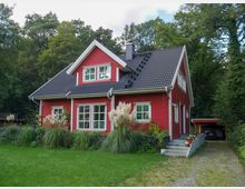 Ein rotes Einfamilienhaus im skandinavischen Stil mit weißen Fenstern und dunkelgrauem Satteldach. Das Haus steht inmitten eines Gartens mit Rasen, Ziergräsern und einer Zufahrt, die zu einem Carport führt, umgeben von dichtem Baumbestand.