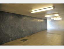 Ein einfacher unterirdischer Durchgang mit gefliesten Wänden und Deckenbeleuchtung durch rechteckige Leuchtstofflampen. Der Boden besteht aus quadratischen Fliesen, und im Hintergrund sind helle Fliesen sichtbar, die den Bereich beleuchten.