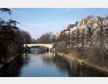 Stadtkanal mit einer Steinbrücke im Hintergrund, gesäumt von kahlen Bäumen auf beiden Seiten und einer Reihe von mehrstöckigen Wohnhäusern auf der rechten Seite. Der Himmel ist klar und blau, das Wasser spiegelt die Umgebung.