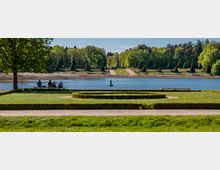 Lakeside park with manicured lawns, low boxwood hedges in geometric beds, and benches along the water’s edge. Across the lake, a tree-lined slope leads to a narrow obelisk-like monument framed by forest.