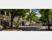 Eine gepflasterte Straße in einer kleinen Altstadt mit Bäumen, die Schatten spenden. Links und rechts sind zweistöckige Gebäude mit Geschäften und Cafés, einige haben Außensitzbereiche mit Sonnenschirmen.