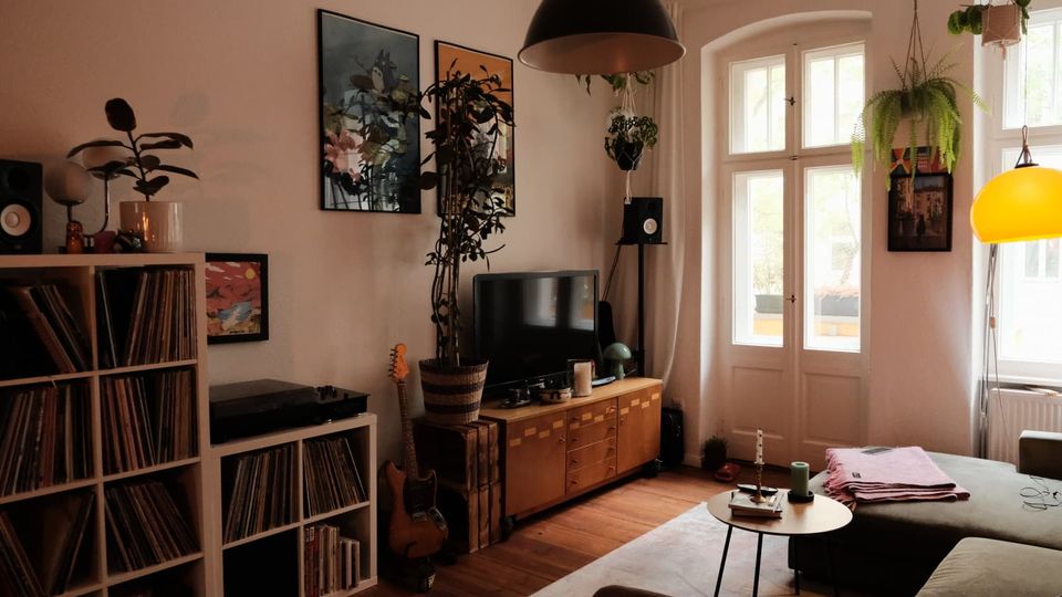 Gemütliches Wohnzimmer mit Holzfußboden, großem Fenster und Glastür, einem TV auf niedrigem Sideboard sowie einem Plattenschrank mit Schallplatten. An den Wänden hängen Bilder, von der Decke hängen Pflanzen und mehrere Pendelleuchten über Sofa und Couchtisch.