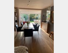 Esszimmer mit großem Holztisch und schwarzen Stühlen auf hellem Holzboden, an das ein Wohnzimmer und ein Garten mit Terrassenmöbeln durch eine Glas-Schiebetür angrenzen. Links ein Bücherregal, rechts eine Kommode und ein an der Wand hängendes Gemälde.