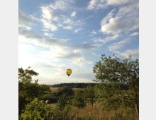 Ein gelber Heißluftballon schwebt über einer ländlichen Landschaft mit Wiesen, Bäumen und einem Heuballen im Vordergrund; im Hintergrund ist eine Baumreihe und ein Himmel mit vereinzelten Wolken zu sehen.