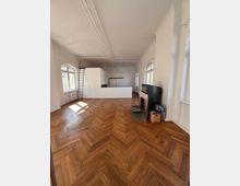 Großzügiger Wohnraum mit Fischgrätparkettboden, weißen Wänden und mehreren hohen Fenstern, die Tageslicht hereinlassen. Im Raum befinden sich ein Kamin, ein kleiner offener Küchenbereich mit weißer Wand, eine Leiter, die zu einer erhöhten Struktur führen könnte, sowie minimalistische Deckenleuchten und sichtbare Rohre.