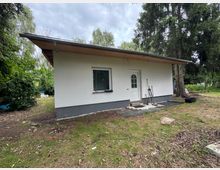 Kleines, weißes eingeschossiges Haus mit flachem Holzdach, einer weißen Tür und einem Fenster. Das Gebäude steht in einer bewaldeten Umgebung mit Wiese und Bäumen im Hintergrund.