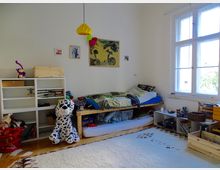 Ein Kinderzimmer mit einem Hochbett, unter dem sich Stauraum und eine Matratze befinden. Links ein weißes Regal mit Büchern und Spielzeug, im Vordergrund ein großer Dalmatiner-Stofftier; rechts ein großes Fenster, das Tageslicht in den Raum lässt.