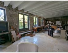 Ein heller Innenraum mit rustikalen Ziegelwänden, großen Fenstern mit Blick auf einen Garten und einer Einrichtung, die auf eine Werkstatt oder ein Atelier hinweist. Der Raum enthält Regale, Tische mit Arbeitsmaterialien und eine Vielzahl von Werkzeugen und Geräten.