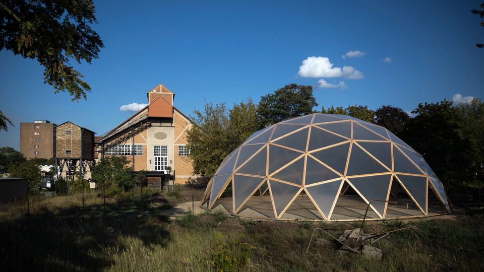 Historisches Industriegebäude aus Backstein mit großen Fenstern und Giebeldach, daneben eine transparente Kuppelstruktur aus Holzstreben auf einer freien Fläche, umgeben von Bäumen und Wiese unter blauem Himmel.