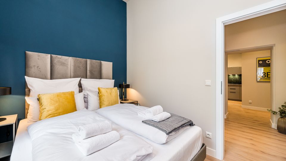 Modernes Schlafzimmer mit Doppelbett, gepolstertem grauen Kopfteil und blau gestrichener Akzentwand; weiße Bettwäsche mit gelben Kissen und Handtüchern auf dem Bett. Rechts führt eine offene Tür in einen Flur mit Holzboden, von dem aus eine Küche zu sehen ist.