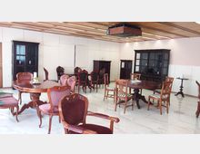 Ein Raum mit eleganter Holzmöblierung, ausgestattet mit runden und rechteckigen Tischen sowie gepolsterten Stühlen im klassischen Stil. Der Boden ist mit glänzendem, weißen Fliesenbelag versehen, die Wände sind schlicht gestaltet, und an der Decke befindet sich eine Holverkleidung mit integrierter Beleuchtung.