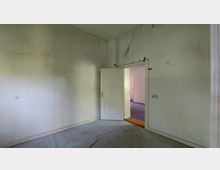 Ein leerer Raum mit hellen, leicht verschmutzten Wänden und einem offenen Türrahmen, durch den ein weiterer Raum mit blauem Teppichboden sichtbar ist. Die Zimmertür ist weiß, und im Raum sind vereinzelt Lichtschalter und Steckdosen erkennbar.