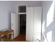 Ein kleines Zimmer mit hellem Holzboden, einer geöffneten weißen Tür und einem hohen weißen Kleiderschrank. Links steht ein kleiner Holztisch mit Büchern und einer gelben Kugel darauf, während Sonnenlicht durch ein Fenster auf die lilafarbenen Wände fällt.