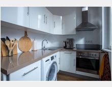 Moderne Einbauküche mit weißen Hochglanzschränken, Edelstahlspüle, Waschmaschine, Cerankochfeld, Dunstabzugshaube und Kaffeevollautomat. Auf der braunen Arbeitsplatte stehen Küchenutensilien in einem Glas, ein Schneidebrett und eine Papierrollenhalterung.