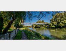 Ein Fußgänger-Steg überquert einen Fluss in einer grünen Parklandschaft mit Bäumen und Sträuchern. Im Vordergrund verläuft ein schmaler Fußweg entlang des Flusses, gesäumt von einem kleinen Zaun und einem überhängenden Baum.