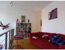 Wohnzimmer mit einer roten Sofaecke und einer weißen Bücherwand an der Rückwand. An den Wänden hängen Kunstbilder und Dekorationen, darunter ein Hirschkopf in Kupferoptik. Ein kleines goldenes Servierwagenmöbel steht vor der Sofaecke auf dem Holzfußboden.