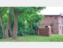 Backsteingebäude mit kleinen Fenstern und einer Mauer im gleichen Stil, umgeben von hohen Bäumen und dichter grüner Vegetation auf einer Rasenfläche. Im linken Bereich des Bildes ist ein schmiedeeisernes Tor erkennbar.