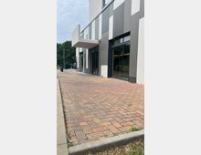 Moderne Fassade eines Gebäudes mit vertikal gestreiften Elementen in Grau- und Weißtönen, großen Glasfenstern und einem hervorspringenden Vordach. Davor ein gepflasterter Gehweg mit verschiedenen Brauntönen und eine Grünfläche am Rand.