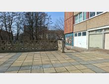 Gepflasterter Vorplatz neben einem mehrstöckigen Gebäude mit Beton- und Klinkerfassade, großen Fensterfronten und einem Keramikmosaik an der Wand. Eine niedrige Beton-Brüstungsmauer mit ornamentalen Öffnungen begrenzt die Fläche; dahinter stehen kahle Bäume und weitere Gebäude im Hintergrund.