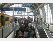 Bahnhofshalle mit modernen Glas- und Metallstrukturen, einer eingefahrenen gelb-roten S-Bahn auf der linken Seite, zahlreichen Reisenden auf Rolltreppen und Treppen sowie Wegweisern zu Ausgängen und S-Bahn-Linien. Im Hintergrund sind große Fenster, Beschilderungen und eine Uhr sichtbar.