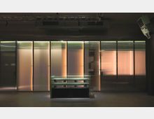 Raum mit moderner Einrichtung und Wänden aus durchscheinenden, strukturierten Paneelen, die von warmem Licht hinterleuchtet sind. Im Vordergrund steht ein Flachmöbel aus Glas und Metallgittern, während oben an der Decke technische Konstruktionen wie Lautsprecher und Traversen sichtbar sind.