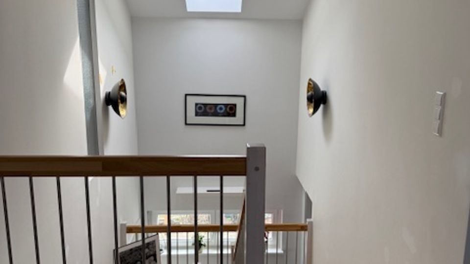 Interior stair landing with white walls and a wooden floor, bordered by a wood handrail and black metal balusters overlooking the staircase below. A rectangular skylight brightens the space, with wall-mounted sconces and framed artwork on the far wall.