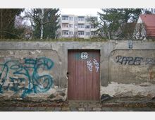 Eine alte Steinmauer mit Graffiti und einer schmalen Holztür in der Mitte. Im Hintergrund sind Bäume und ein mehrstöckiges Wohngebäude zu sehen.