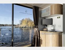 Innenansicht eines modernen Hausboots mit einer kleinen Küchenzeile oder Pantry, bestehend aus einem Spülbecken, Hängeschrank und Fernseher. Durch eine große Glastür ist eine Aussicht auf einen Pier und einen ruhigen See mit kahlen Bäumen im Hintergrund zu sehen.