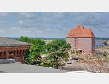 Backsteingebäude mit steilem Ziegeldach auf freier Fläche, umgeben von Bäumen, mit Blick auf einen Fluss und angrenzender Landschaft im Hintergrund. Im Vordergrund ein detailreicher Ausschnitt eines weiteren Ziegelgebäudes und Zweige.
