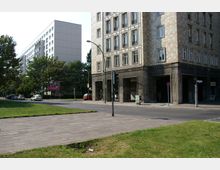 Straßenszene mit einem mehrstöckigen Gebäude aus Steinfliesen rechts im Bild und einer Wohnanlage mit heller Fassade links im Hintergrund. Im Vordergrund sind eine Grünfläche, ein gepflasterter Gehweg und eine Straße mit parkenden Autos und einer Ampel zu sehen.