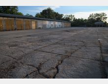 Großer leerer Betonplatz mit Rissen und Abnutzungsspuren, umgeben von einem flachen Gebäude mit gelben Toren sowie Bäumen im Hintergrund. Die Szene wirkt ländlich oder industriell, teils im Schatten und vom Sonnenlicht beleuchtet.