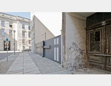 Straßenszene mit einem breiten Gehweg aus Steinplatten, flankiert von einem modernen Gebäude mit großem Eingang und einer schlichten Metalltoranlage rechts. Im Vordergrund steht eine alte, verwitterte Holztür mit Gitterverzierungen und einer beschädigten Fassade mit Graffiti.