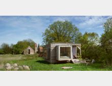 Kleines, modernes Holzhaus mit großen Fenstern steht auf einer grünen Wiese, umgeben von Bäumen und Sträuchern. Im Hintergrund sind ein historisches Steingebäude und der Turm einer Kirche sichtbar, die den ländlichen Charakter der Umgebung betonen.