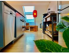 Moderne Wohnküche mit Holzboden, ausgestattet mit Edelstahlgeräten wie Geschirrspülmaschine und Backofen auf der linken Seite sowie einem Holzregal mit Küchenutensilien rechts. Im Hintergrund befindet sich ein Dachfenster mit natürlichem Lichteinfall und eine rote Stehlampe nahe einem geflochtenen Möbelstück.